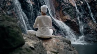 Beyaz elbiseli genç bir kadın bir kayanın üzerinde şelalenin arka planında oturuyor ve yoga yapıyor.
