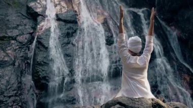 Beyaz elbiseli genç bir kadın bir kayanın üzerinde şelalenin arka planında oturuyor ve yoga yapıyor.