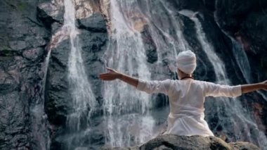 Beyaz elbiseli genç bir kadın bir kayanın üzerinde şelalenin arka planında oturuyor ve yoga yapıyor.