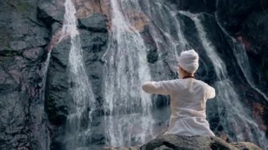Beyaz elbiseli genç bir kadın bir kayanın üzerinde şelalenin arka planında oturuyor ve yoga yapıyor.