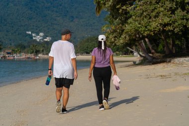 Yaşlı, çok uluslu bir çift yaz sabahı sahilde sağlıklı bir yürüyüş yapıyor.