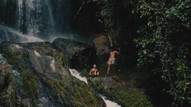 Genç bir çift kayalara tırmanıyor ve güneşli bir yaz gününde tropiklerde bir şelaleye bakıyor.