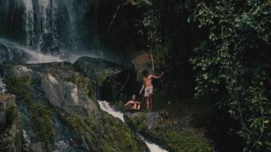 Genç bir çift kayalara tırmanıyor ve güneşli bir yaz gününde tropiklerde bir şelaleye bakıyor.