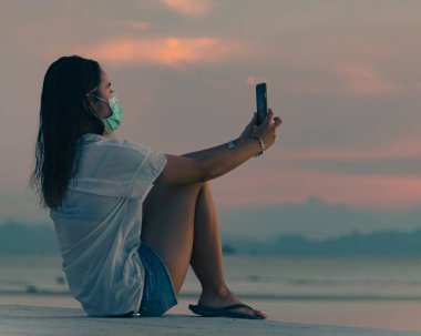 Elinde akıllı telefonuyla maske takan genç bir kadın setin üzerinde oturur ve denizin üzerinde günbatımının tadını çıkarır.