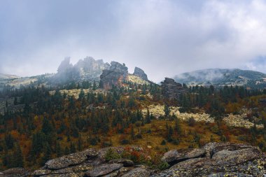 Sonbaharda ağaçlar ve kayalarla kaplı bir dağ vadisi manzarası. Bulutlar, sis ve sis dağın tepesinde..