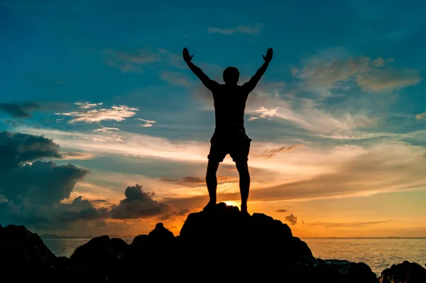 Щасливий чоловік стоїть на скелі тропічного острова з руками вгору і спостерігає за заходом сонця — стокове фото
