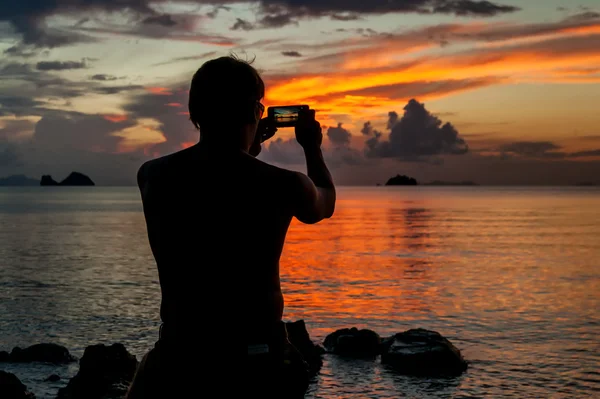 男子站在岸边的一个热带岛屿和拍摄日落您的电话 — 图库照片