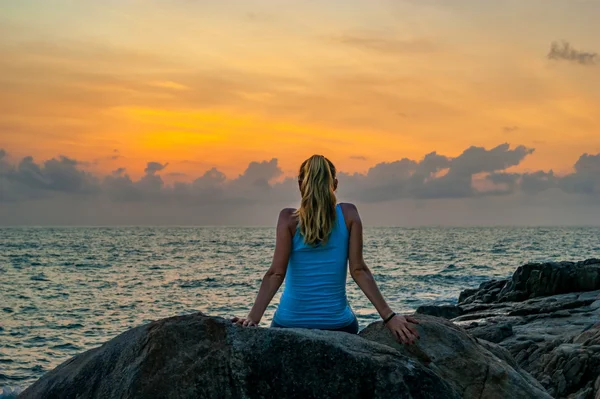 Young woman sitting on the rocks by the sea and watching the sunrise on a tropical island — Stock Photo, Image