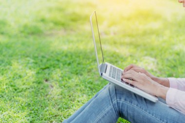 Asyalı kadın yeşil parkta dizüstü bilgisayar kullanarak oturuyor. Dizüstü bilgisayarlı mutlu girişimci işi yapan kadın Coronavirus karantina döneminde elleri klavye ev ofisinde yazan bir not defteri kullanıyor.