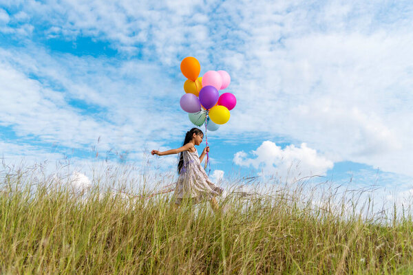 Cheerful cute girl holding balloons running on green meadow white cloud and blue sky with happiness. Hands holding vibrant air balloons play on birthday party happy times summer on sunlight outdoor