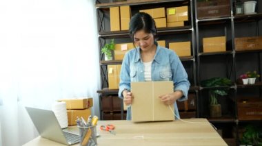Kadın paket kutusunu masaya koydu. Müşteriye mal teslimatını kontrol ediyor. Asyalı bir kadın ofis masasında küçük bir iş kuruyor. Girişimci Asyalı kadın teslimat için ürün topluyor