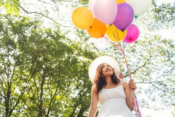 Cheerful beauty woman holding balloons relax sitting under big tree in green park with happiness. Woman Hands holding vibrant air balloons play on birthday party happy time summer on sunshine outdoor