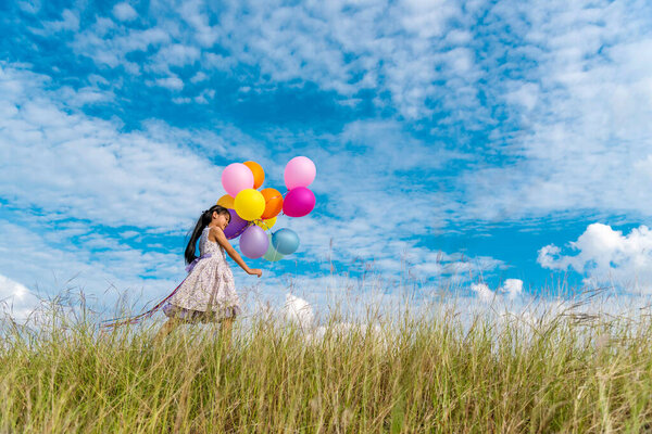 Cheerful cute girl holding balloons running on green meadow white cloud and blue sky with happiness. Hands holding vibrant air balloons play on birthday party happy times summer on sunlight outdoor