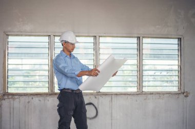 İnşaat mühendisi planı tutuyor. İnşaat alanında çalışan işçilerden mavi gömlekli, güvenlik önleyici beyaz şapka giyiyor. Mimarlık mühendisi, inşaat mühendisi, emlak konseptine bakıyor.
