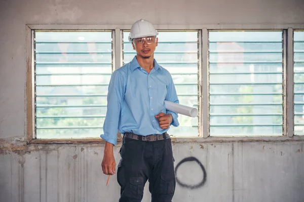 建設エンジニアの男性は 建設現場の労働者で青いシャツの安全性の白いハード帽子を着用青写真を保持します 建築家の技術者土木労働者は青写真不動産の概念を見る — ストック写真