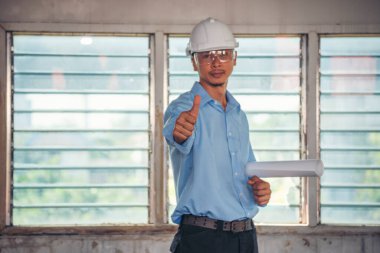 İnşaat mühendisi planı tutuyor. İnşaat alanında çalışan işçilerden mavi gömlekli, güvenlik önleyici beyaz şapka giyiyor. Mimarlık mühendisi, inşaat mühendisi, emlak konseptine bakıyor.