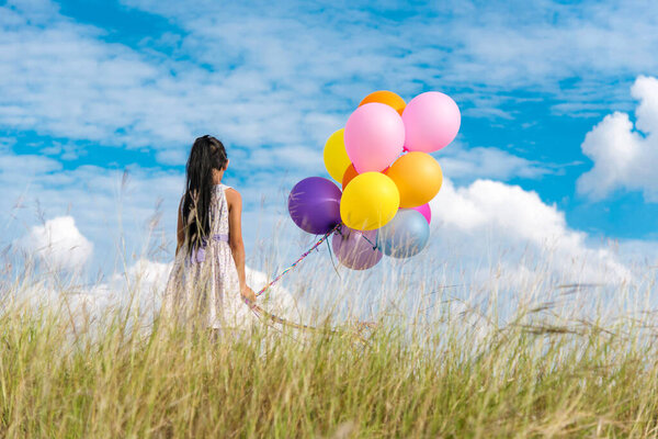 Cheerful cute girl holding balloons running on green meadow white cloud and blue sky with happiness. Hands holding vibrant air balloons play on birthday party happy times summer on sunlight outdoor