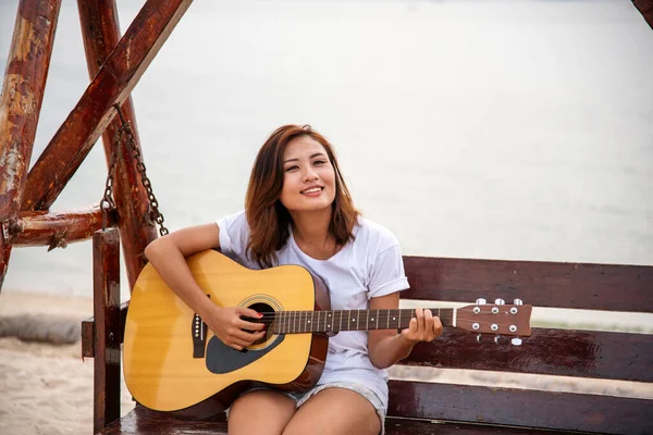 Genç kadınlar sahilde akustik gitar çalıyorlar. Kum tropik plaj adasında oturan mutlu kadın akustik gitar enstrümanı çalıyor. Asyalı müzisyen kız Mutlu gitarist hobisi