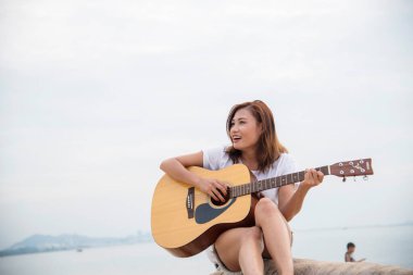 Genç kadınlar sahilde akustik gitar çalıyorlar. Kum tropik plaj adasında oturan mutlu kadın akustik gitar enstrümanı çalıyor. Asyalı müzisyen kız Mutlu gitarist hobisi