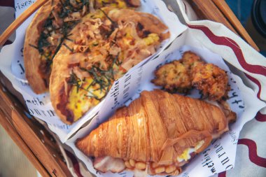 Piknik sepeti kruvasan ekmeği pastırmalı yumurta peynirli brunch, yiyecek kutusu kahve fincanı portakal suyu kırmızı battaniye üzerinde ahşap hasır kutuda. Buğday ekmeği, fırın, meyve, yiyecek ve yeşil park, açık hava yaşam tarzı.