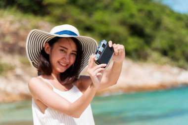 Yaz aylarında mavi gökyüzünde dijital kamera kullanan bir kadın gezgin fotoğraf çekiyor. Asyalı Fotoğrafçı Kadınlar yaz aylarında plajda rahatlar. Mutlu gülümseyen kadın fotoğraf çekerken okyanus adasının tadını çıkar