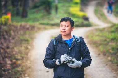 Asyalı adam fotoğraf çekmek için profesyonel dijital kamera kullanıyor. Gülümseyen erkek fotoğrafçı dışarıdaki fotoğraf makinesine bakıyor. Yeşil doğa parkında fotoğraf çeken yetişkin bir adam. Güzel doğa erkekleri hobisi