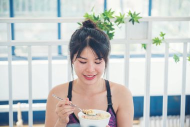 Süt organik besin maddeleriyle yulaf ezmesi yiyen Asyalı kadınlar. Kadınlar ellerinde kaşık ve kase ile sağlıklı süt ürünleri mısır gevreği, meyve ve muz tutuyorlar. Evde sağlıklı beslenme tarzı