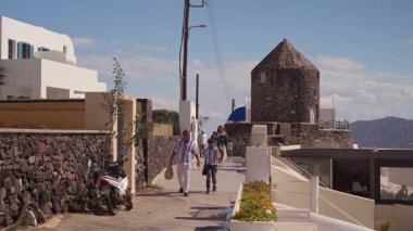 Santorini, Yunanistan 'da Imerovigli - 7 Ekim 2025 - İnsanlar güneşli bir günde eski bir taş yel değirmeninin yanında yürüyorlar.