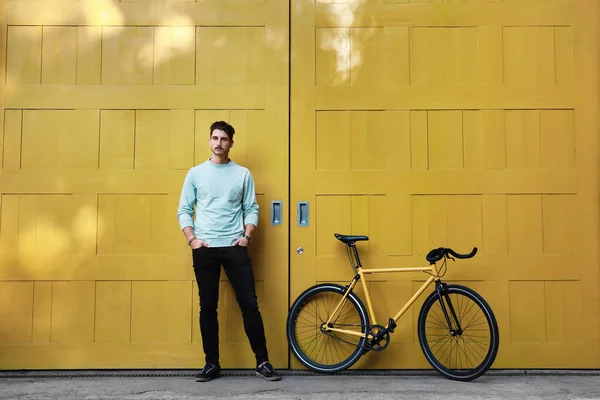 Um jovem elegante homem caucasiano hipster posando ao lado de sua bicicleta no fundo amarelo. — Fotografia de Stock