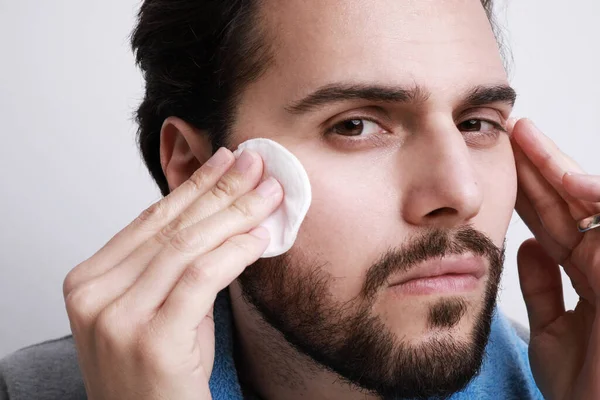 Bearded man with the help of a hygienic cotton pad is cleaning his face skin.