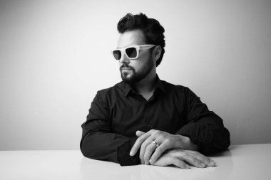 Sakallı adamın beyaz güneş gözlüğü taktığı siyah beyaz bir fotoğraf. Stüdyo oturumu.