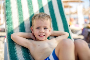 4 yaşında çizgili şortlu bir çocuk kumsalda güneşli bir yatakta uzanıyor, mutlu ve mutlu. Yaz, deniz kıyısı