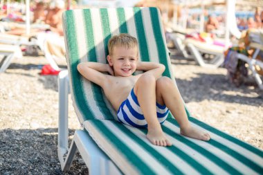 4 yaşında çizgili şortlu bir çocuk kumsalda güneşli bir yatakta uzanıyor, mutlu ve mutlu. Yaz, deniz kıyısı