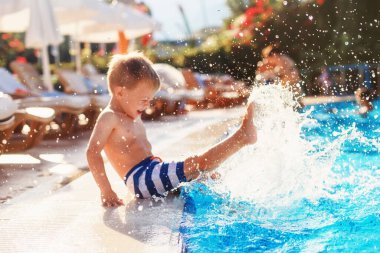 Çizgili şortlu küçük bir çocuk otelin yanındaki havuza ayaklarını vuruyor, burnunu güneşten kırıştırıyor. Yaz tatili. Seçici odak
