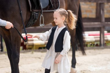 6-7 yaşlarında bir kız bir atı okşuyor ve atı dizginleyerek onu beslemek istiyor. Çocuklar için binicilik sporu