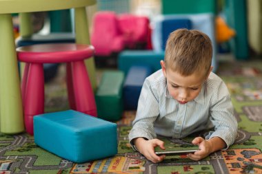 6-7 yaşlarında bir çocuk oyun odasında halıya uzanıyor ve telefonda oynuyor. Çocukları ve aletleri kavrayın