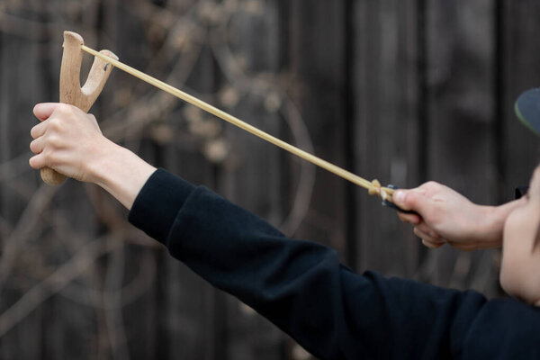 A boy in a black jacket with a hood and a cap shoots from a slingshot, blurred background