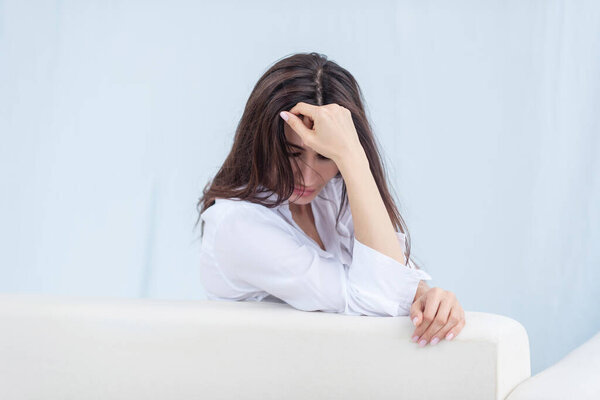 Брюнетка склонила голову на руку и изображает печаль, недоумение, сидит на светлом диване на белом фоне