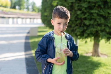 7-8 yaşlarında bir çocuk parkta plastik bir bardaktan limonata içiyor ve kameraya bakıyor.