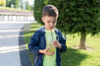 7-8 yaşlarında bir çocuk parkta plastik bir bardaktan limonata içiyor ve kameraya bakıyor.