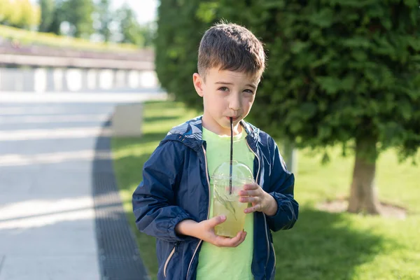 7-8 yaşlarında bir çocuk parkta plastik bir bardaktan limonata içiyor ve kameraya bakıyor.