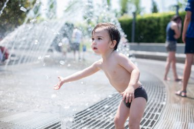 Çekici küçük bir çocuk çeşmeye su sıçratıyor, yüzünde bir gülümseme ve haz. Şehirde yaz konsepti