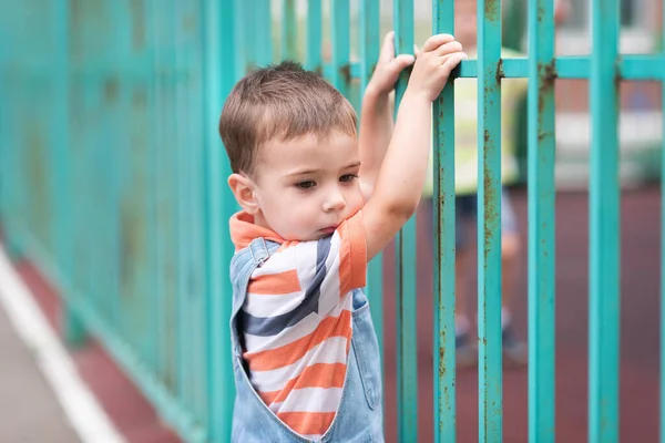 A boy on the playground, a tall green fence, a fencing, with peeling paint.