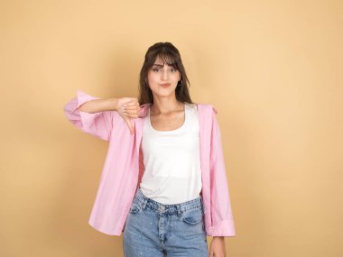 Hayal kırıklığına uğramış üzgün kadın, 20 'li yaşlarda genç beyaz esmer kadın hayal kırıklığına uğramış üzgün kadın sıradan pembe tişört beyaz tişört giyiyor bej arka planda izole edilmiş baş parmak sevmeme hareketi yapıyor, stüdyo portresi çiziyor..