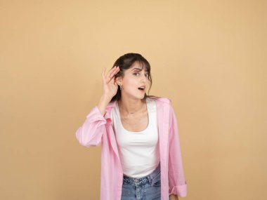Pembe gömlekli meraklı genç bir kadın elini kulağına dayayıp dikkatlice dinlemek için öne eğiliyor. Tarafsız bir bej arka plana karşı duruyor. Şaşırmış ve meraklı bir yüz ifadesi var..