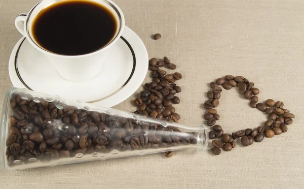 Coffee Cup and saucer on linen fabric. Coffee beans in heart shape glass bottle — Stock Photo, Image