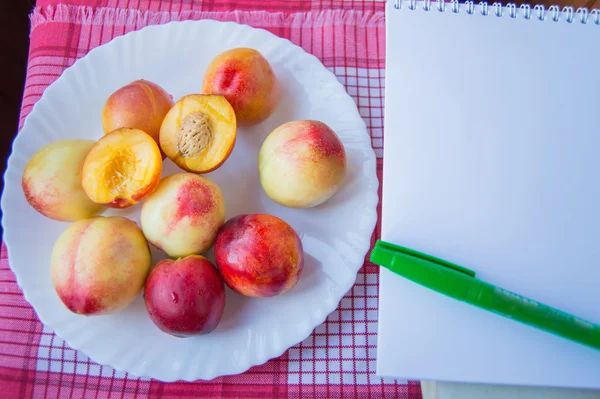 Спелые персики на белой тарелке с блокнотом и ручкой — стоковое фото