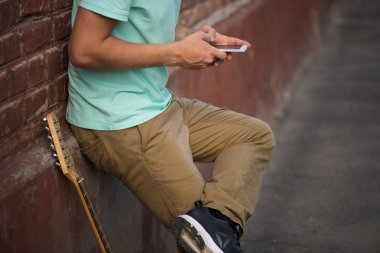 Genç şık adam kahverengi tuğla duvar, cep telefonu yanında gitar holding eller olmalıdır