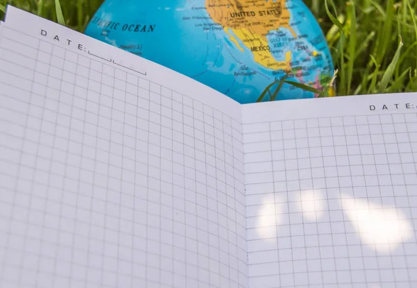 An open empty notebook lies on the grass near the globe, the concept of ecology and back to school, school summer holidays, a place for text.