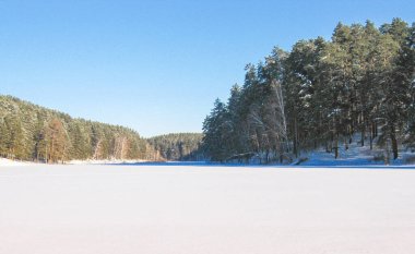 Buz ve karla kaplı donmuş bir gölün kış manzarası, kozalaklı ormanlarla çevrili, güneşli soğuk bir gün..